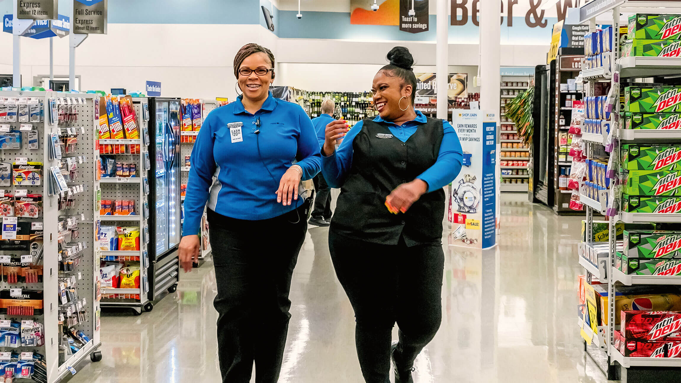 Two women working in a supermarket smiling and talking to each other whilst walking toward the camera