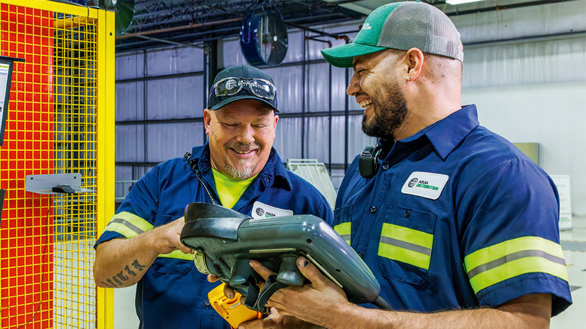Two warehouse workers in blue uniforms with reflective stripes smiling and operating a handheld device near yellow safety barriers.