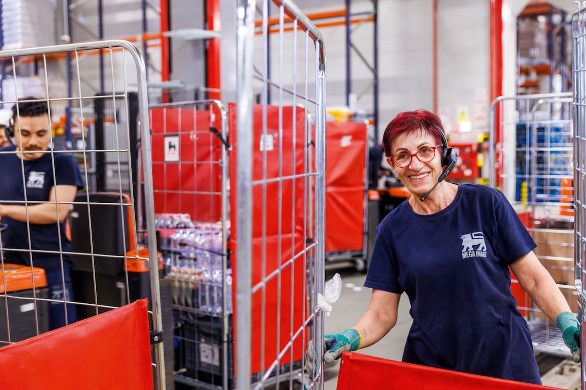 Smiling female warehouse worker with headset and gloves standing by a metal cart in a store or distribution center.