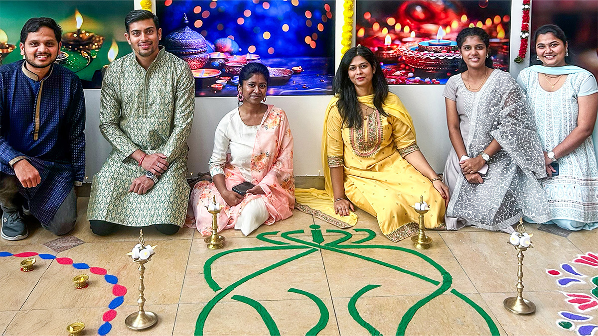 Six people dressed in traditional Indian attire sitting on the floor behind colorful rangoli designs and brass oil lamps, with a festive background featuring lit candles.