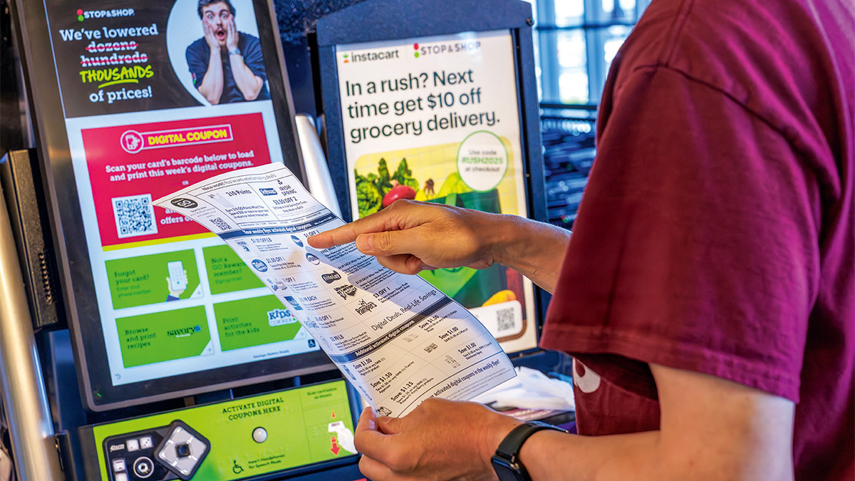 Person holding and pointing at a printed coupon sheet in front of a self-service digital coupon kiosk.