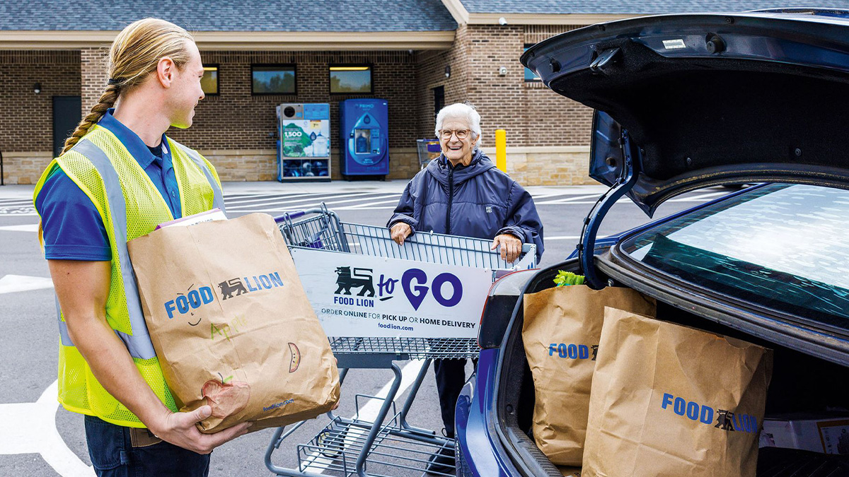 Young male employee in safety vest holding grocery bags loading an elderly woman's car in a parking lot with shopping cart nearby.